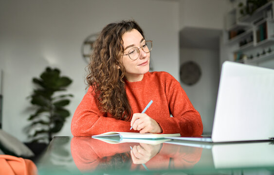 Young student woman student using laptop elearning, remote working at home office looking at computer watching webinar, virtual learning, having video conference call hybrid meeting, writing notes.