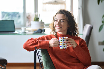Happy calm lady enjoying warm hot drink with mug in hands daydreaming in the morning. Young adult smiling pretty woman sitting on chair holding cup drinking tea or coffee relaxing at home looking away