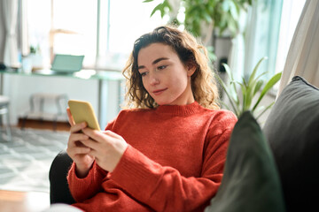 Relaxed young woman sitting on couch using cell phone technology, happy lady holding smartphone,...