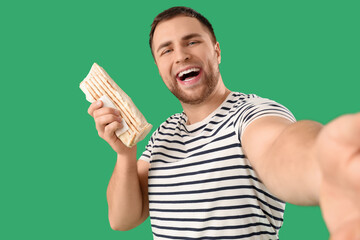 Handsome young man with tasty doner kebab taking selfie on green background