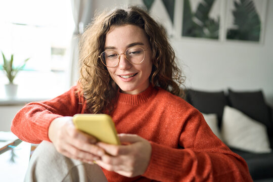 Happy girl checking cell phone online education apps, texting, browsing internet for shopping sitting at home. Young smiling woman wearing glasses holding smartphone using cellphone, looking at mobile - Powered by Adobe