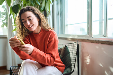Happy young woman sitting on chair holding mobile phone using cellphone device, looking at...