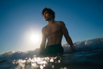 Fit man with wet tousled hair emerging from deep blue ocean wearing sporty green swim trunks at sunrise