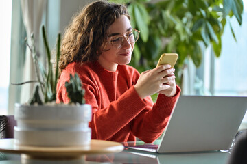 Happy young lady using smartphone social media apps at home. Smiling woman holding cellphone modern...