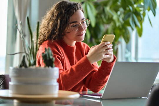 Happy young lady using smartphone social media apps at home. Smiling woman holding cellphone modern ai applications, looking at mobile, checking cell phone, texting chatting sitting at table at home. - Powered by Adobe