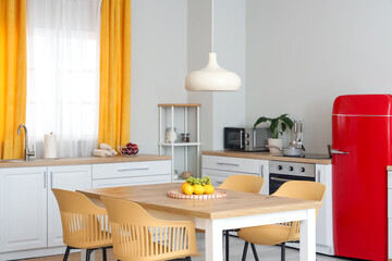 Interior of modern kitchen with white counters, dining table, red fridge, chairs and lamp