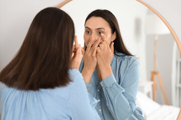 Young woman putting in contact lenses near mirror at home