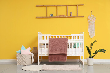 Interior of stylish children's bedroom with crib, cozy blanket and houseplant near yellow wall