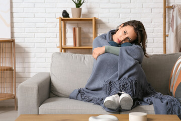 Depressed young woman wrapped in plaid on sofa in living room