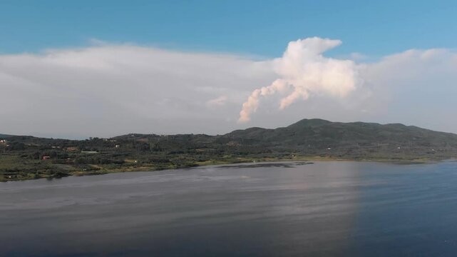 Scenic aerial footage capturing the serene landscape of Corfu Island, featuring a calm body of water (potentially a lake or bay) reflecting the sky, surrounded by lush green mountains under a dynamic 