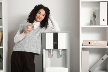 Young businesswoman with glass of water near cooler in office