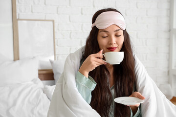 Beautiful young woman with sleep mask, blanket and cup of coffee sitting in bed at home