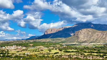 Fototapeta premium The stunning view of majestic mountains and lush fields stretches beneath a bright blue sky with fluffy clouds