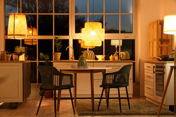 Interior of modern kitchen with counters, dining table and glowing lamps in evening