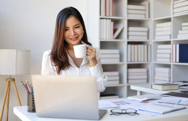 Happy Asian businesswoman in the office drinking hot coffee and working with laptop at the desk in the morning.