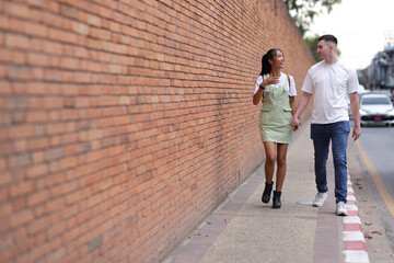 Couple of tourists walking and holding hands on a roadside sidewalk in a tourist attraction area in a city in Thailand. Fellow tourists are walking in a crowded community.