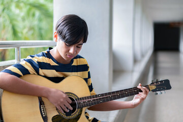 Teen boy playing acoustic guitar, wearing a yellow shirt, looking down to guitar thoughtfully. Creative youth expression and musical lifestyle in a casual setting.