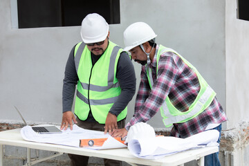 Asian overweight foreman working on construction site wearing safety white helmet consult contractor male middle-aged partnership, two Asian worker inspects blueprint paperwork using laptop technology