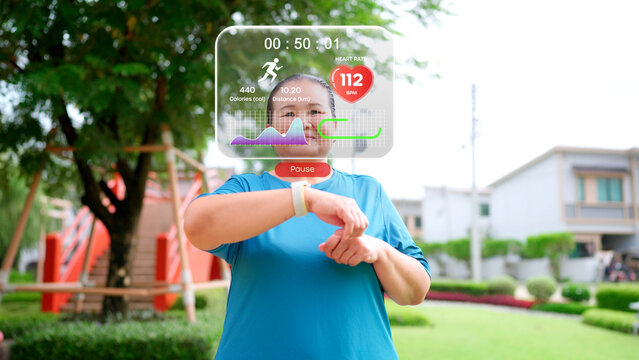 Active senior Asian woman checking fitness data on a smartwatch with a futuristic augmented reality interface while jogging in a park, representing modern health technology and a healthy lifestyle.
