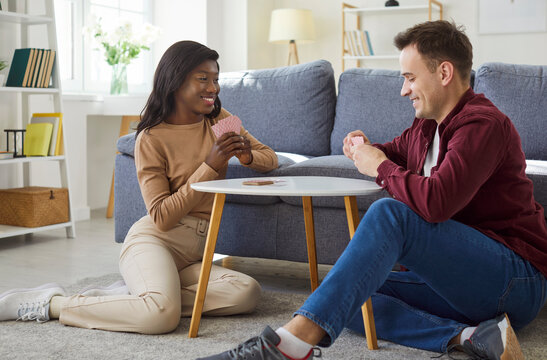 Beautiful young multinational couple in love having fun playing card games sitting on floor in living room at home. Happy man and woman relaxing and enjoying leisure time together.