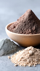 Three distinct piles of powdered clay in gray, beige, and brown shades displayed on a textured surface, with the brown powder in a wooden bowl.