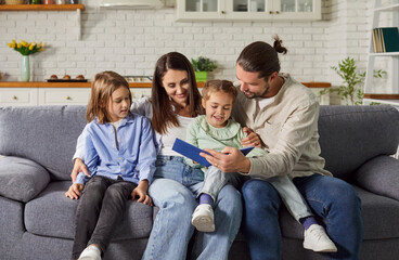 Young happy family reading a book with their little cute daughter and son sitting on sofa at home. Smiling parents spending time with children and smiling. Leisure time together concept.