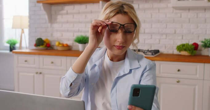 Portrait of a young woman in eyeglasses suffering from presbyopia sitting in kitchen at home trying to read message on smartphone. Farsightedness eye disease and vision problems. 4k video.