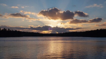 Obraz premium Serene lake view at sunset with sun rays piercing through the clouds and reflecting on the water surface.
