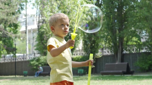 A young boy is focused on blowing a large bubble with a yellow bubble wand. He is playing outdoors in a sunny, green park in Simferopol, Crimea.