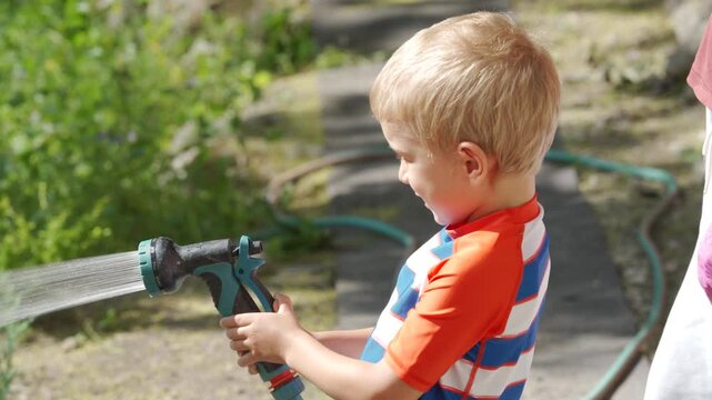 A young boy holds and sprays water from a garden hose. He is enjoying a bright sunny day outdoors in Simferopol, Crimea, surrounded by greenery.