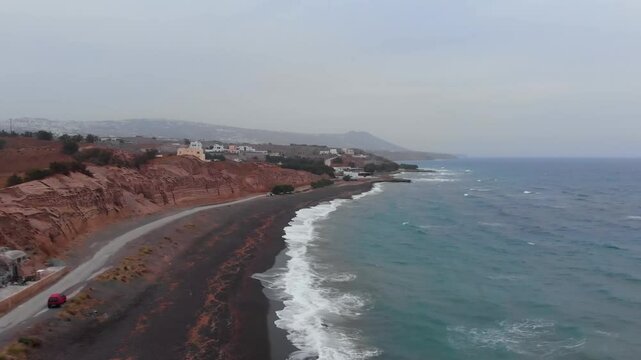 An aerial shot captures the unique beauty of Santorini's Red Beach (Kokkini Paralia), showcasing its distinctive red cliffs, dark volcanic sand, and the clear turquoise waters of the Aegean Sea.