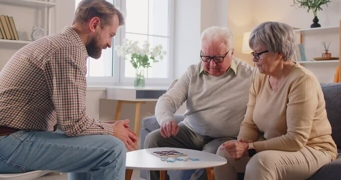 Smiling middle-aged man playing board game with elderly parents at small table in living room. Happy senior couple enjoying leisure time, bonding with adult son, engaging in fun family activity.