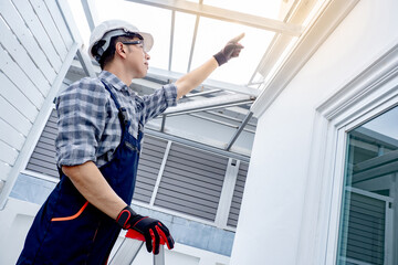 Asian male foreman or construction worker climbing up on an aluminium ladder pointing at the roof structure of a new house. Professional builder working at housing project building site