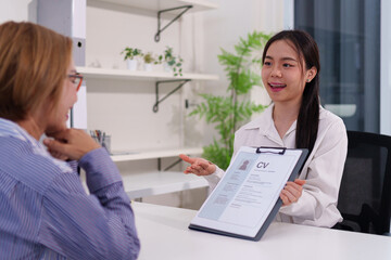 Asian Young Woman Interviewing Adult Candidate with Resume CV on Clipboard in Office Setting Discussing Job Opportunities and Qualifications