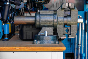 Metal vise mounted on a wooden workbench in a workshop. Tools and a blue perforated panel are...