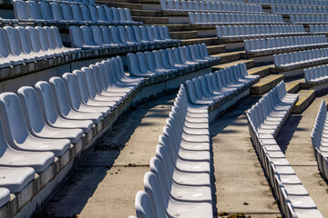 Naklejka premium Empty plastic seats arranged in rows on a concrete stadium grandstand, illuminated by bright sunlight.