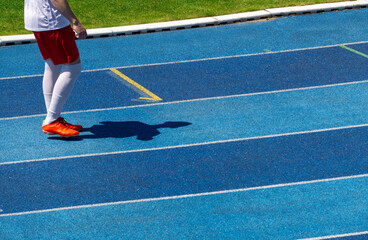 Football player in red shorts and white socks standing on a blue athletics track. Bright colors and a dynamic shadow emphasize the sporty atmosphere of the scene.