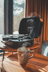 Close-up of a steaming coffee mug on wooden table with a vinyl record player in the background,...