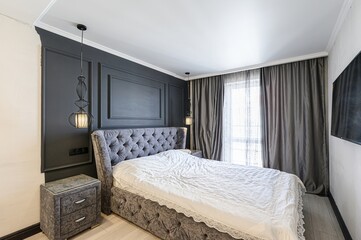 stylish bedroom featuring a tufted grey bed, dark accent wall, and modern pendant lights. Curtains...