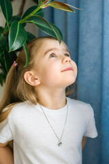 Smiling Little Girl in White Clothes with a Necklace Posing Near Houseplant, Looking at the Camera.