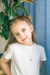 Smiling Little Girl in White Clothes with a Necklace Posing Near Houseplant, Looking at the Camera.