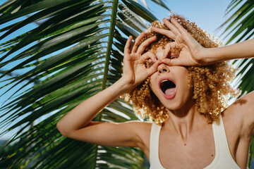 playful woman with curly hair making binoculars gesture under palm leaves, joyful expression, sunny...