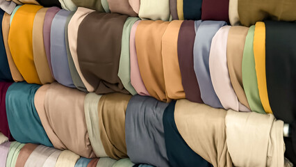 A stack of rolls of headscarf fabric in various earthy and muted colors. The texture and color palette of the textiles provide a rich and warm visual aesthetic