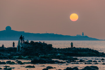 Tourists gather at the rocky shore during spring sunset to admire and photograph the tranquil ocean...