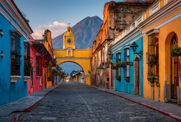 Obraz premium Cityscape of the colorful main street of Antigua city at sunrise with the famous yellow arch and the Agua volcano in the background, Guatemala, Central America.