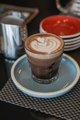 A glass of layered coffee beverage topped with intricate latte art featuring a heart design