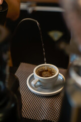 A close-up shows a thin stream of liquid pouring from a curved spout into a white ceramic cup on a...
