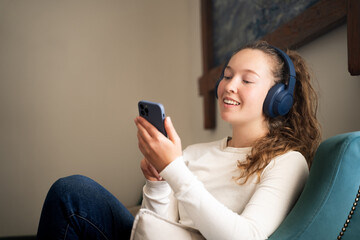 Young woman using mobile phone smiling wearing wireless headphones sitting on sofa in living room...