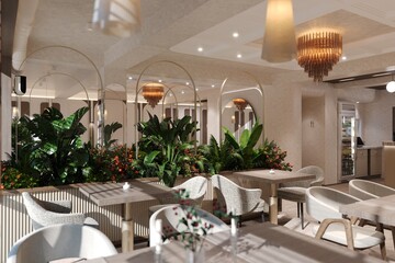 Beautiful modern café interior with lush indoor plants, elegant arch mirrors, and warm natural light. The space features textured seating, refined chandeliers, and a cozy, inviting atmosphere, blendin