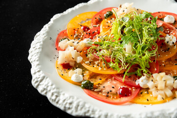 Colorful fresh salad with sliced tomatoes, diced cheese, greens, and seeds on a white plate over black background. Healthy vegetarian meal, close-up.